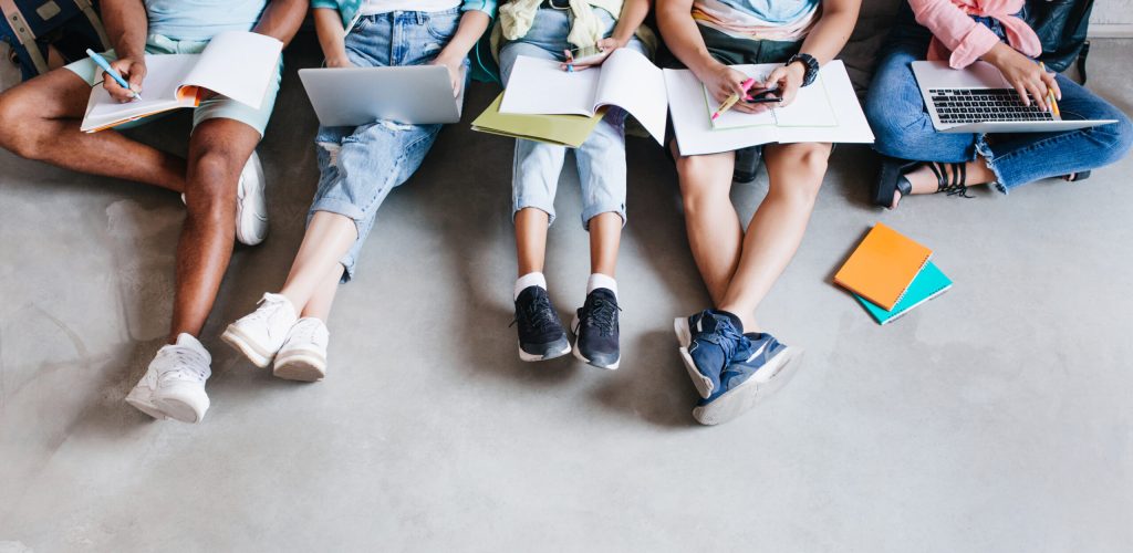 Overhead portrait of young people with laptops and smartphones, sitting together on the floor. Students writing lectures holding textbooks on their knees.